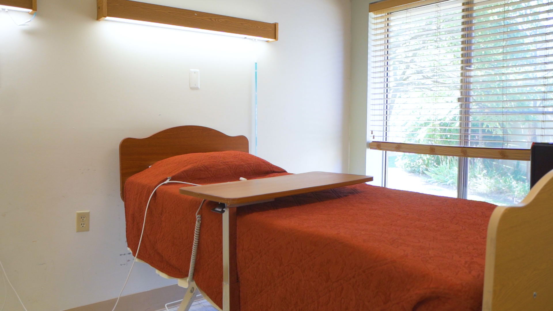 Room with one bed and orange blanket with bed table. One window on the background.